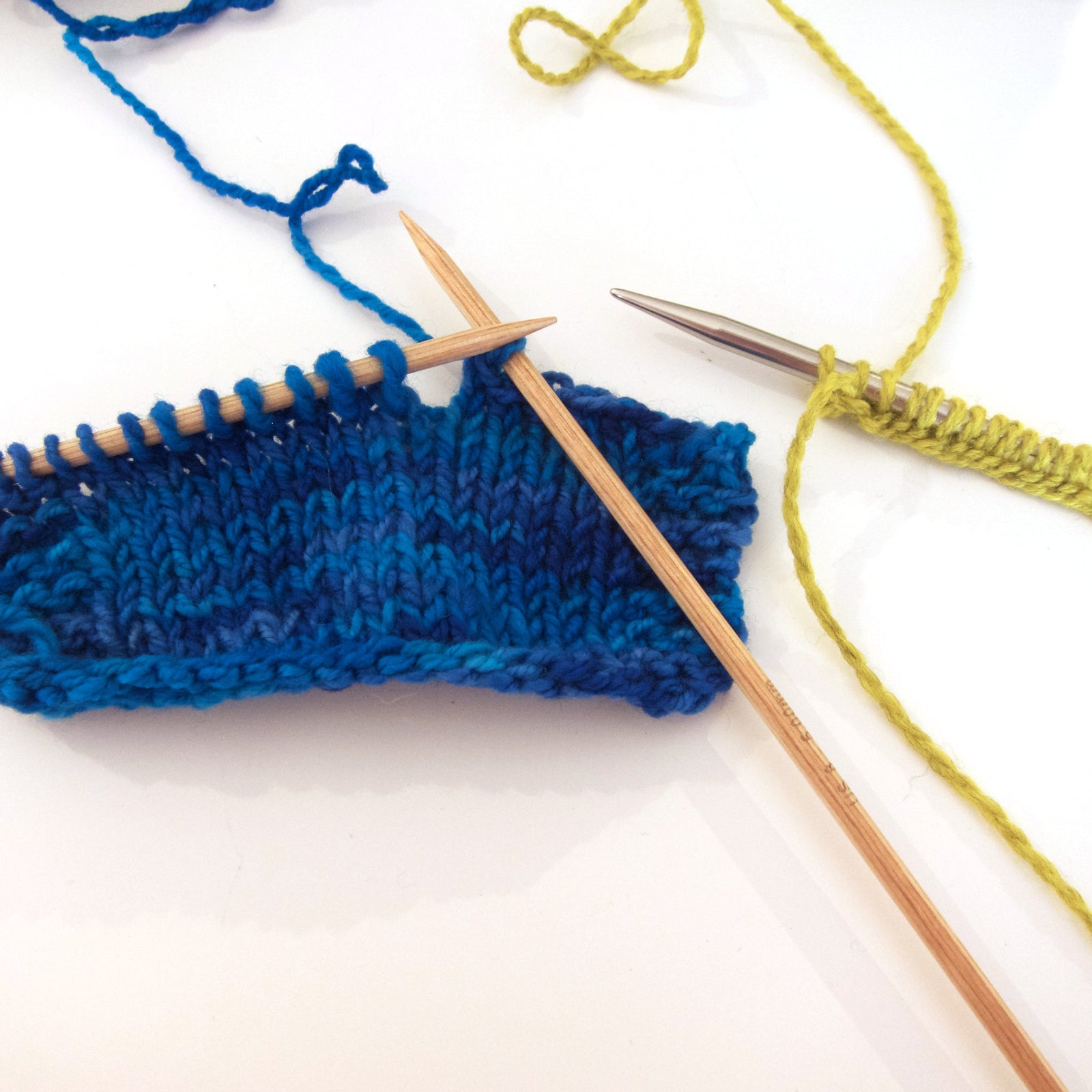 Knitting needles and knitting.  Blue yarn knit on wooden straight needles and bright green yarn on metal needles. 