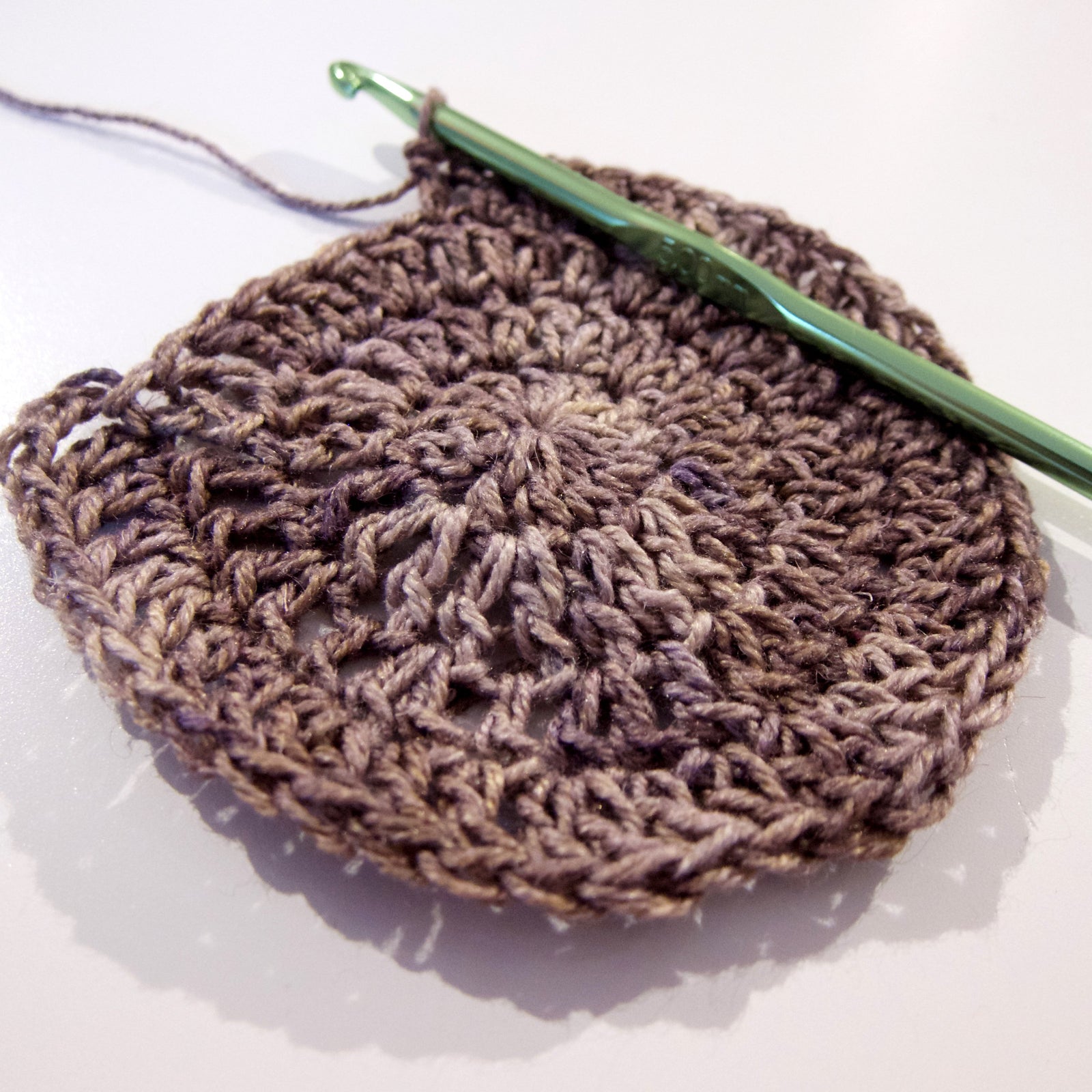 A green crochet hook resting on top of a grey-brown circle of crochet stitches