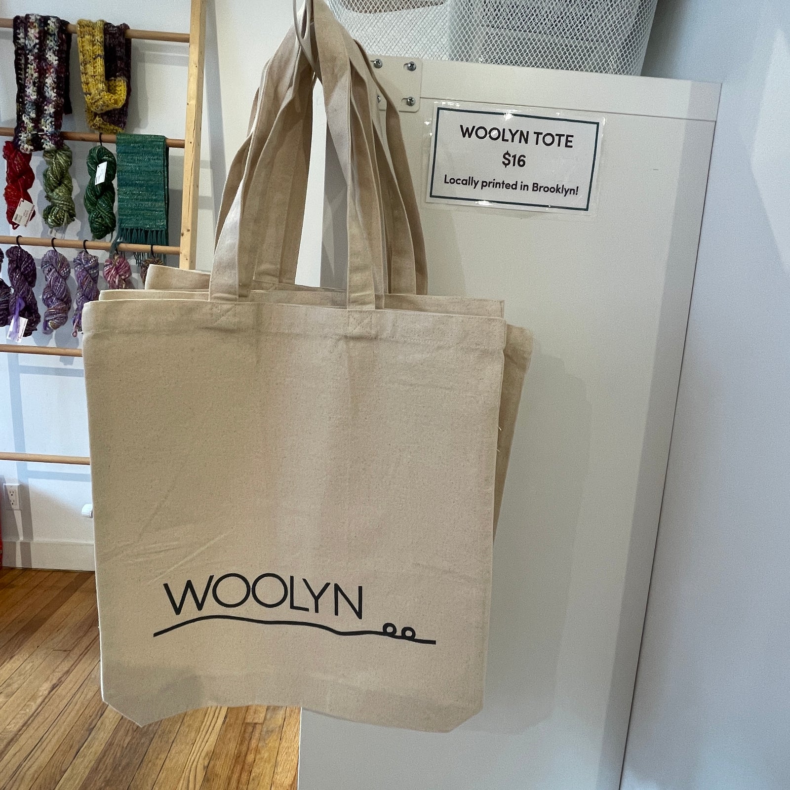 Image of a canvas tote bag hanging on the edge of a shelf. It features the Woolyn name and logo printed at the bottom in black ink.