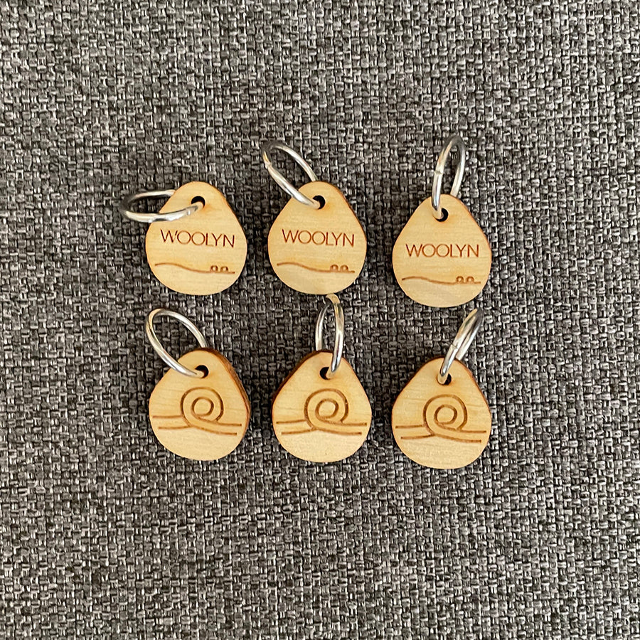 Image of a set of six wooden stitch markers with metal rings. The stitch markers are teardrop shaped and feature the Woolyn name and logo.