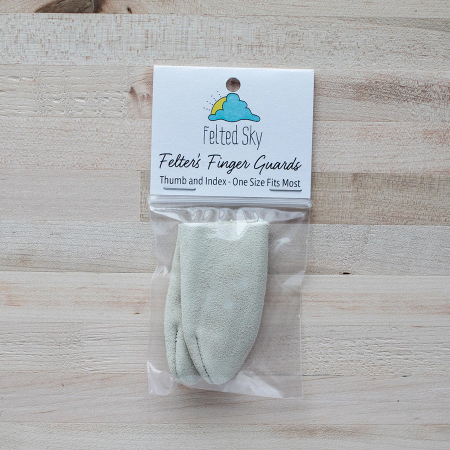 A pair of white suede finger guards in a plastic package with white paper label displayed on a pale wood background