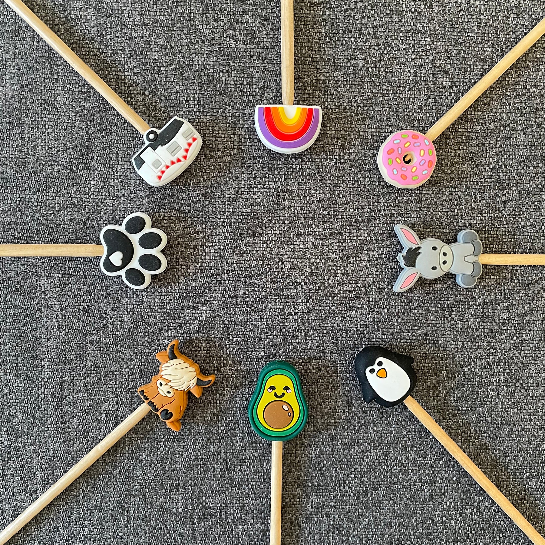 Knitting needles topped with rainbow, donut, donkey, penguin, avocado, cow, paw print, and camper Stitch Stoppers arranged in a circle
