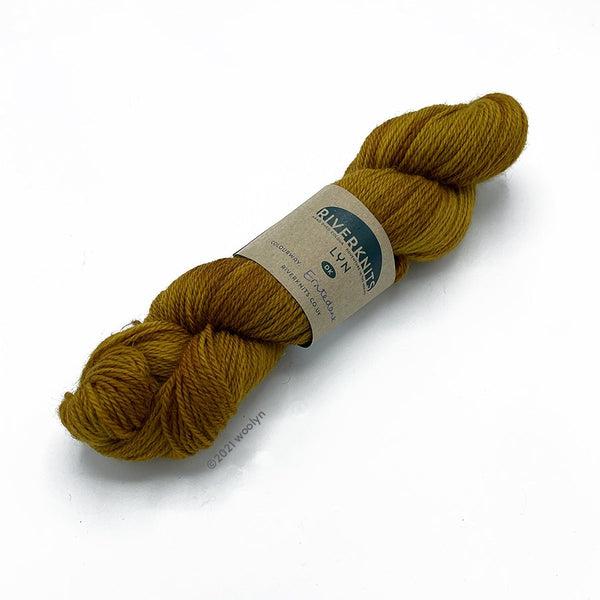 A skein of River Knits Lyn a worsted plied DK weight yarn in Eintedank a mustard color with earthy brown overtones.
