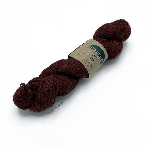 A skein of River Knits Lyn a worsted plied DK weight yarn in Kastaine, a dark maroon with chesnut undertones.