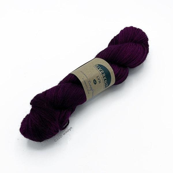 A skein of River Knits Lyn a worsted plied DK weight yarn in Zwergpflaume, a deep purple color.
