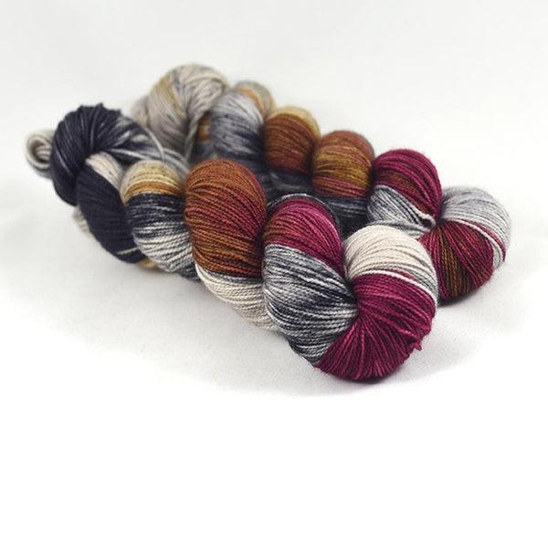 Skeins of Destination Yarn Passport Palais Garnier, a variegated yarn with deep crimson, warm golden brown, hints of warm yellow and several shades of grey ranging from light to dark. 
