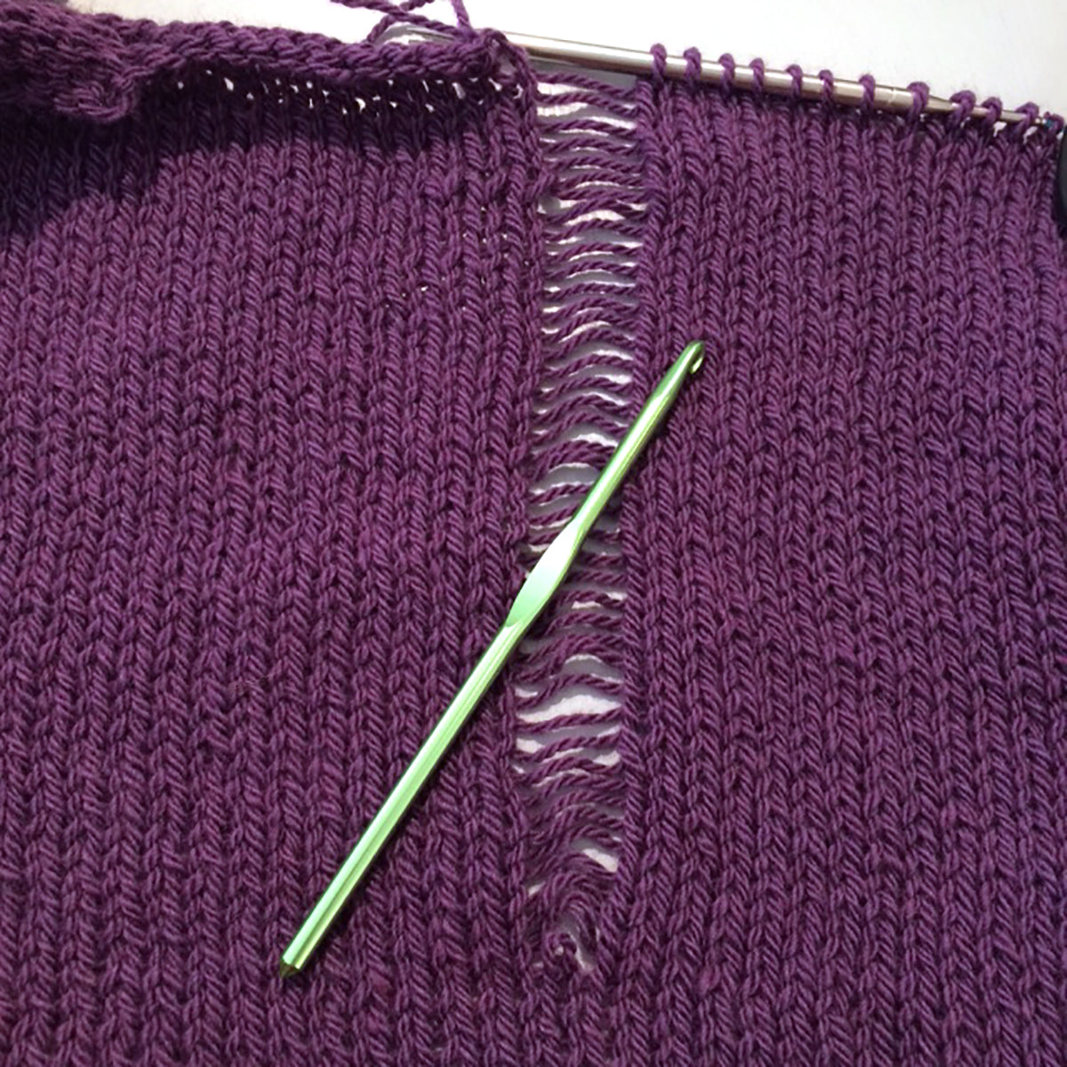 Green crochet hook lying atop purple knitting where one stitch has dropped down. 