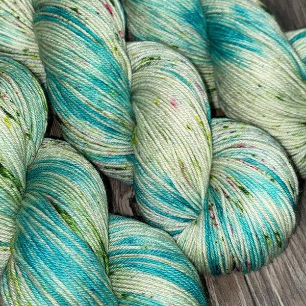 Skeins of JEMS Monster Minis Persephone, a variegated and speckled yarn in shades of pale green and bright turquoise with red and dark green speckles.