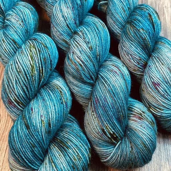 Skeins of JEMS Monster Minis Queen of the Underworld, a variegated yarn in grey blues and teal with purple and green speckles. 