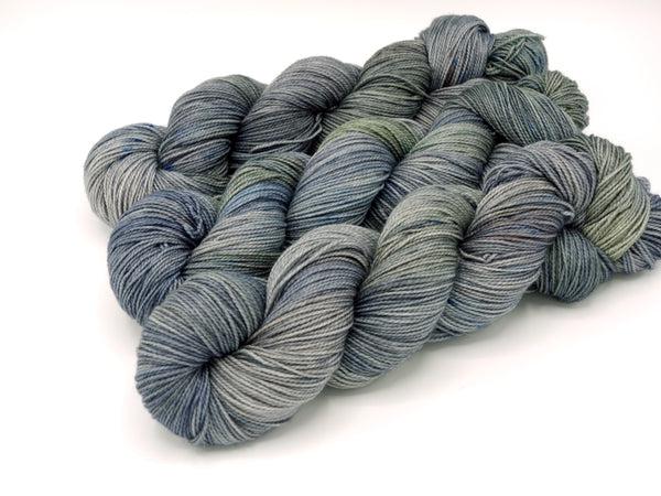Skeins of Murky Depths Deep Sock Tempest, a gently variegated yarn with shades of grey. green grey and hints of dark purple. 